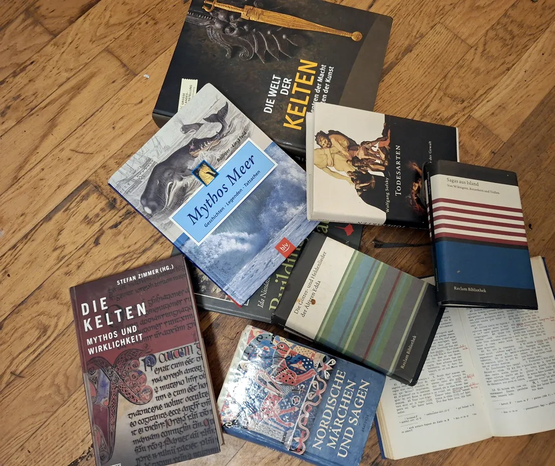Bücher über Kelten, Wikinger, nordische Sagen und das Meer auf einem Holzboden, darunter die Edda und ein aufgeschlagenes Buch mit roten Randnotizen